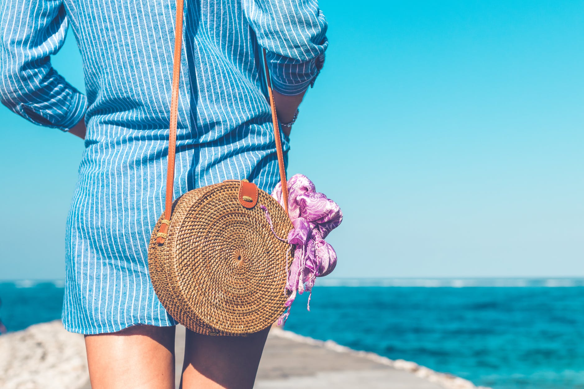 woman wearing blue and white striped dress with brown rattan crossbody bag near ocean